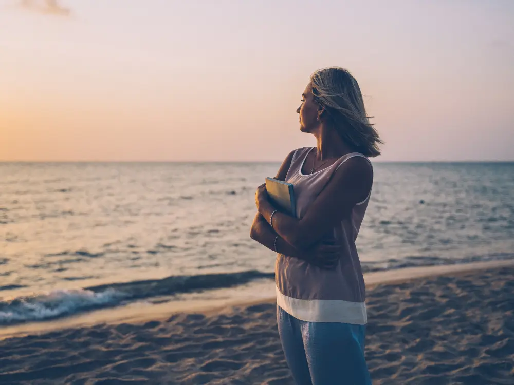 Mulher loira na praia ao pôr do sol, abraçando um livro e olhando para o mar, representando a reflexão sobre a síndrome do ninho vazio aos 40 anos.